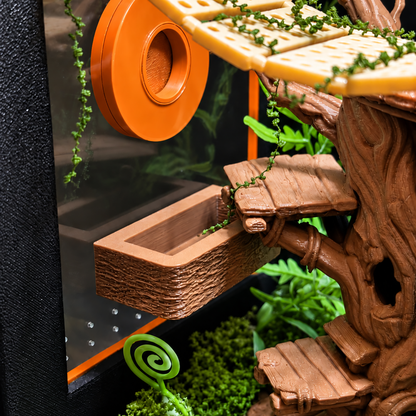 A close-up of a small treehouse-style pet play structure with faux wood textures, green vine accents, a yellow platform, and an orange circular wheel or tunnel piece against a dark background.