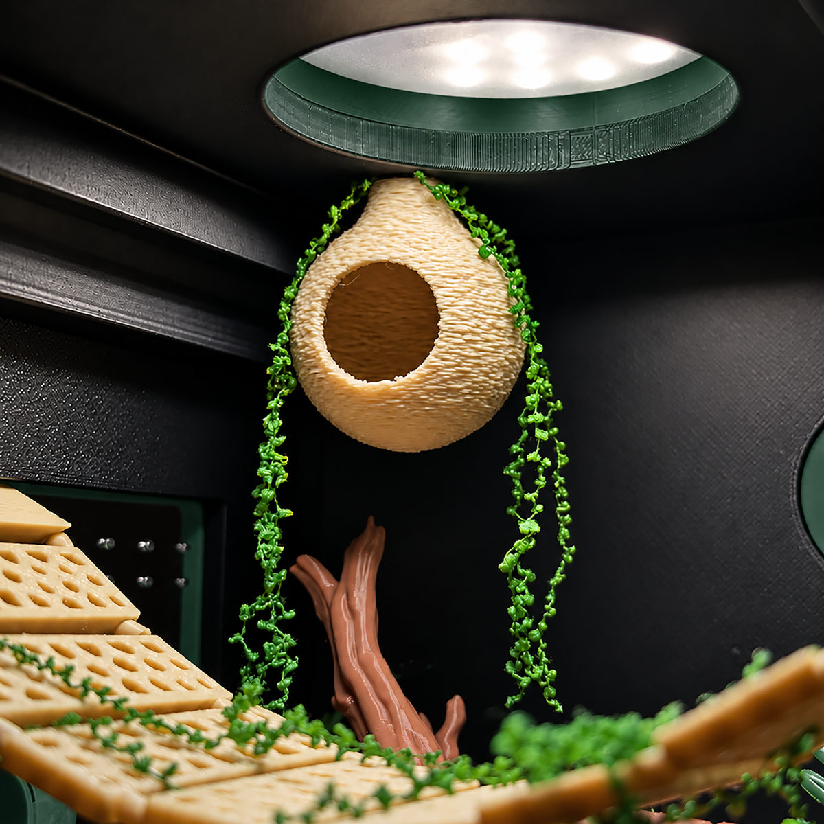 A close-up of a 3D-printed beehive nest with hanging green vines and a wooden branch inside a dark enclosure, under a green light dome.