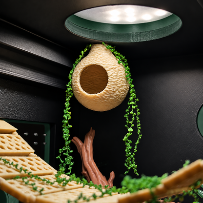A close-up of a 3D-printed beehive nest with hanging green vines and a wooden branch inside a dark enclosure, under a green light dome.
