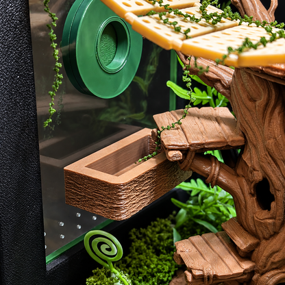 A close-up of a terrarium or aquarium interior featuring a brown faux-wood tree trunk structure with branches, a yellow elevated platform, green hanging vines, moss, and a green circular filter or decoration attached to the glass.