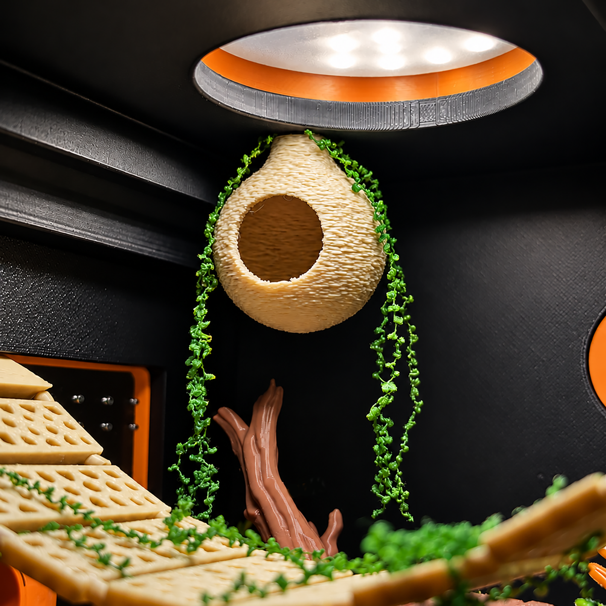 A close-up of a miniature diorama or playset interior with a beige woven hanging nest, green vine decorations, brown faux logs, and tan platform pieces against a dark black backdrop with orange accents.