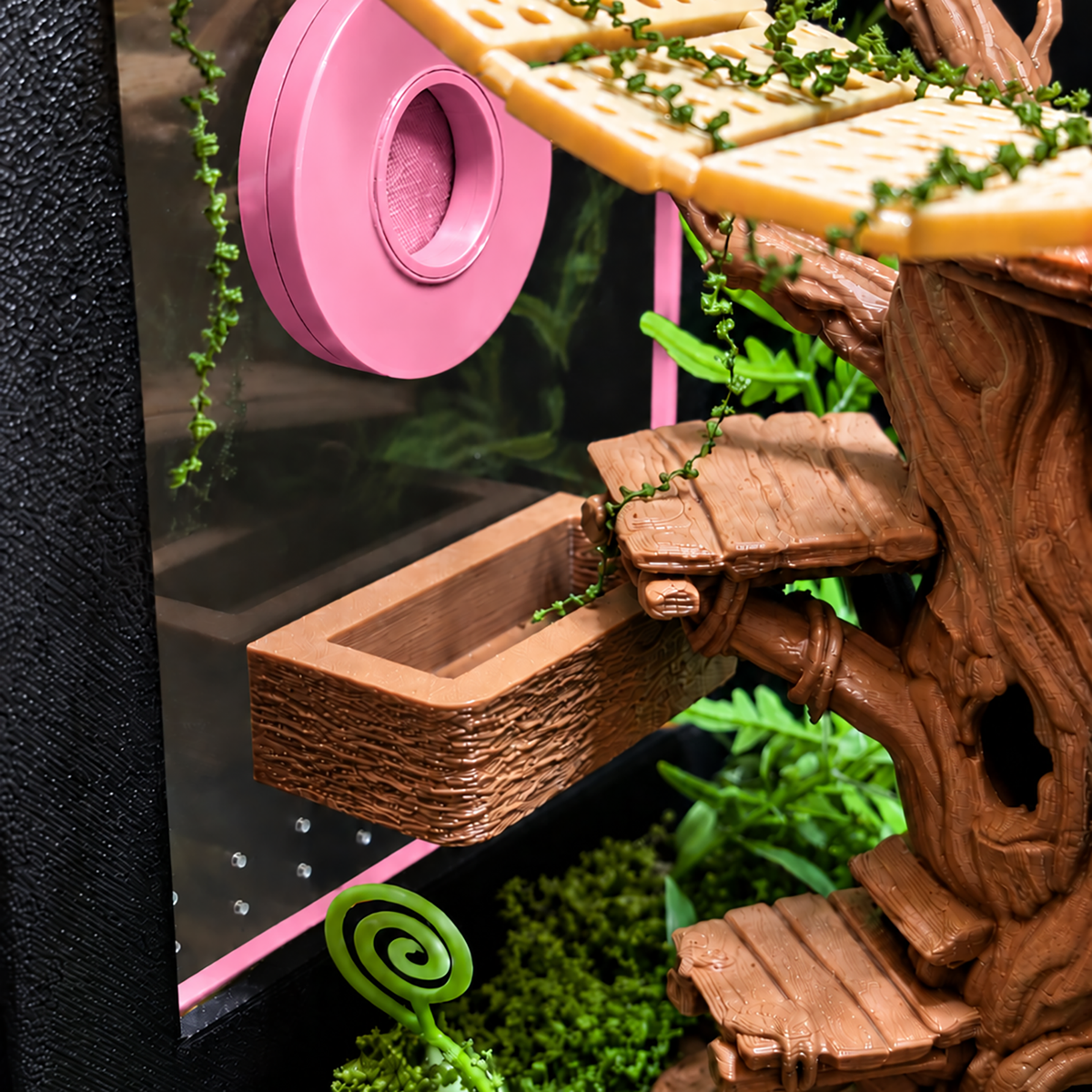 A close-up of a small treehouse-style terrarium accessory with brown faux-wood planks, green vine details, a pink circular wheel, and a yellow platform inside a black and pink enclosure.