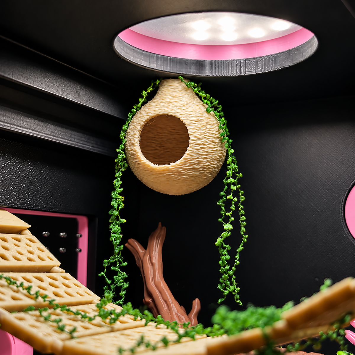A close-up of a 3D-printed animal hide or enclosure interior, featuring a hanging textured beige hide, green faux-ivy vines, wooden branches, and curved beige platforms against a black background with pink accents.